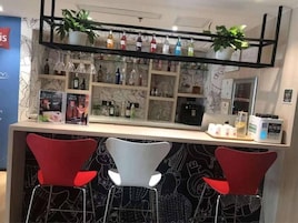 Zona de bar con dos sillas rojas, estanterías con botellas y una planta.