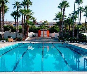 A swimming pool with clear blue water, surrounded by palm trees and a building in the background. - Kartibubbo Beach Resort (Campobello di Mazara)