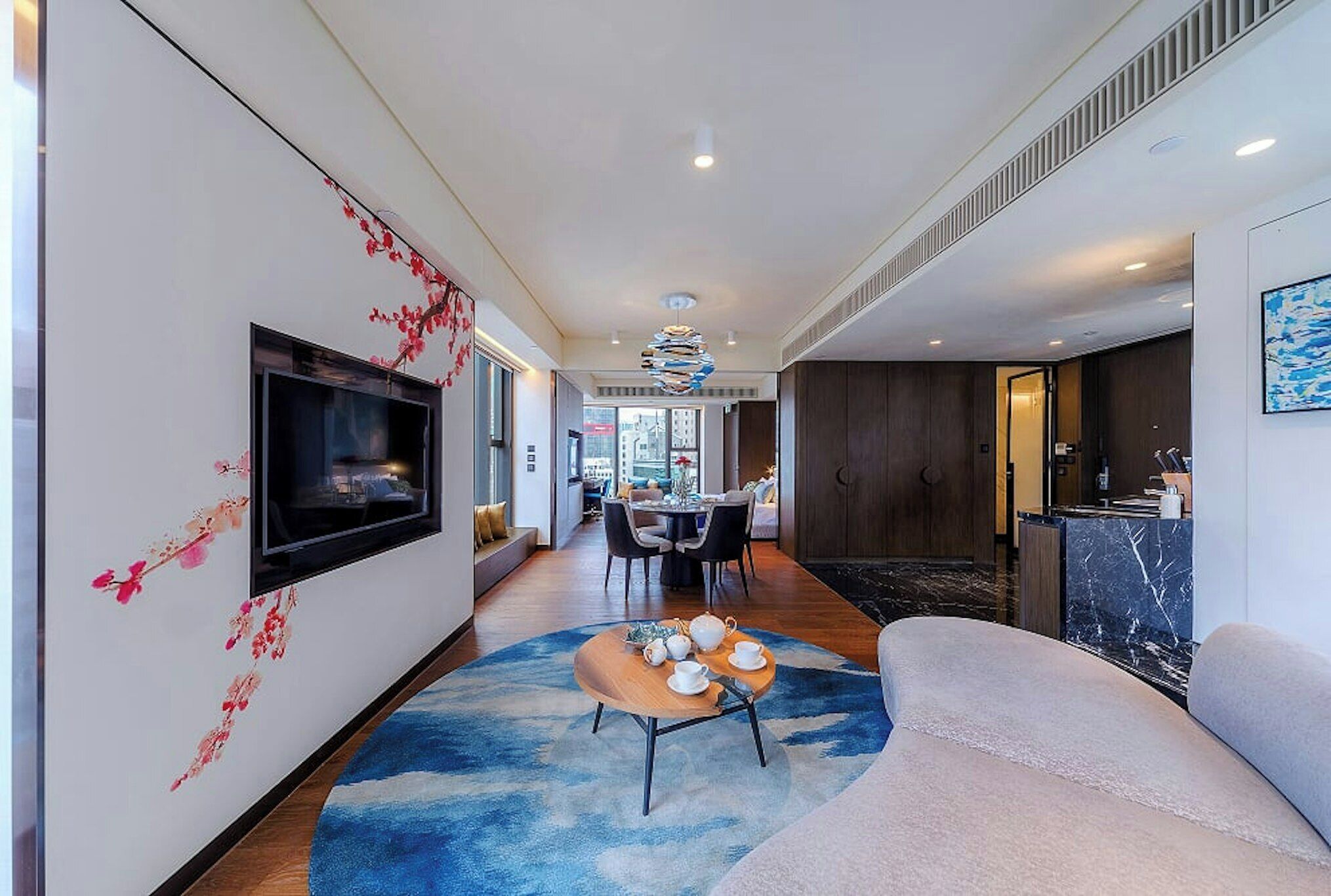 A modern living room with a flat-screen TV, a round coffee table with teapots, a sofa, and a dining area with chairs and a table.