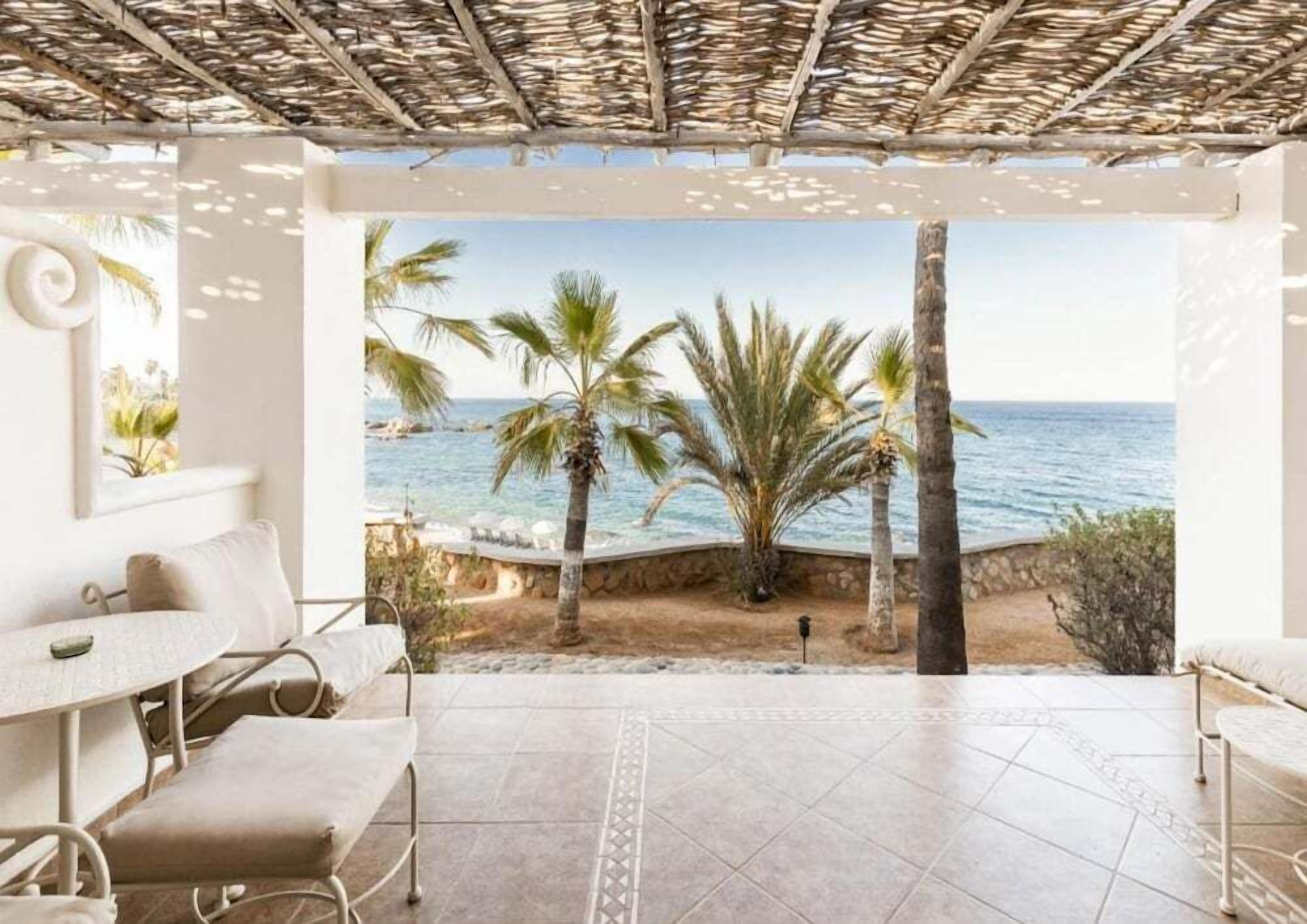 A covered outdoor seating area with a view of the sea and palm trees.