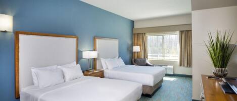A hotel room with two beds, a desk, a chair, a plant, and a window with curtains.
