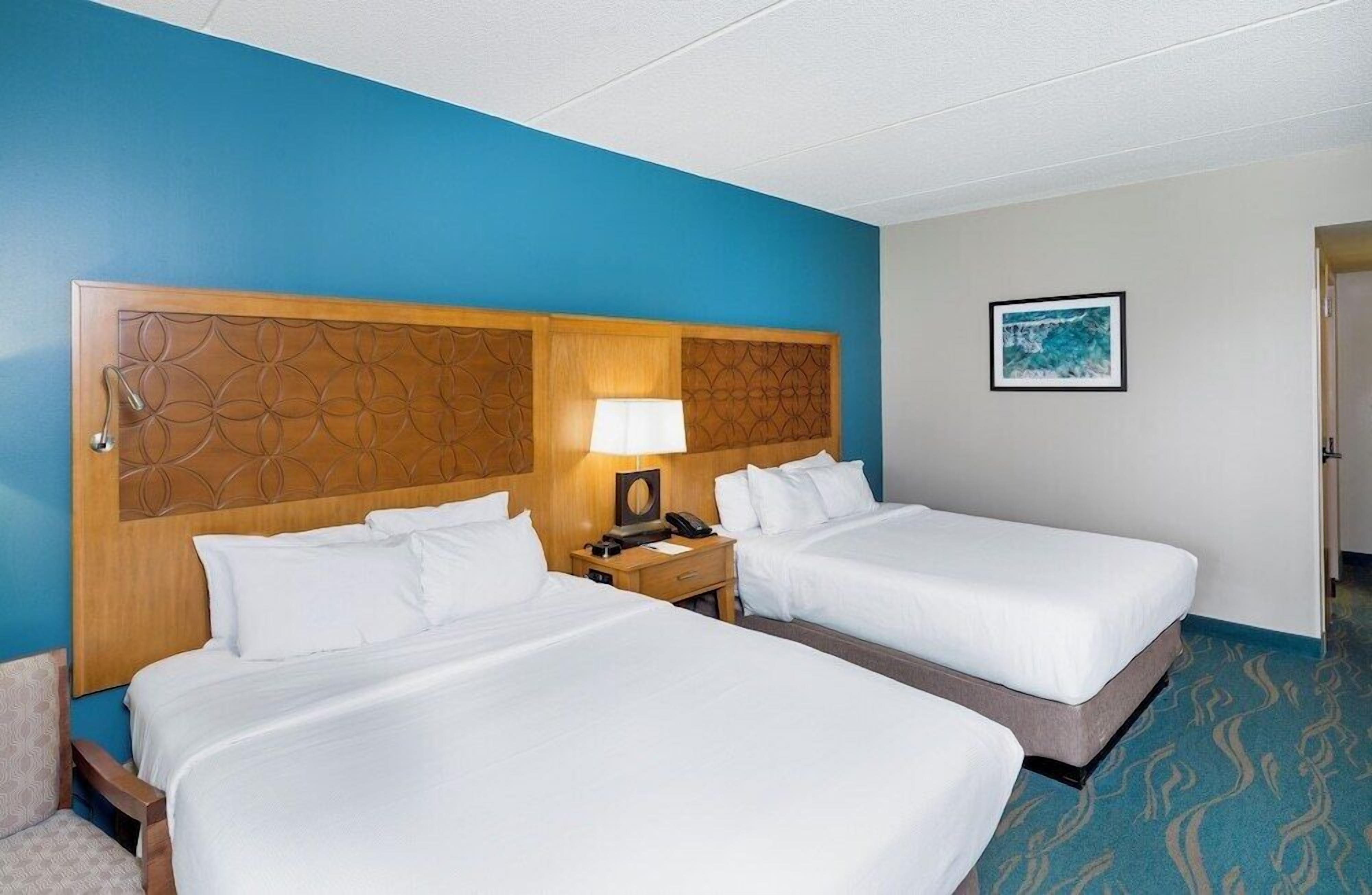 A hotel room with two beds, a wooden headboard, a nightstand with a lamp, and a framed picture on the wall.