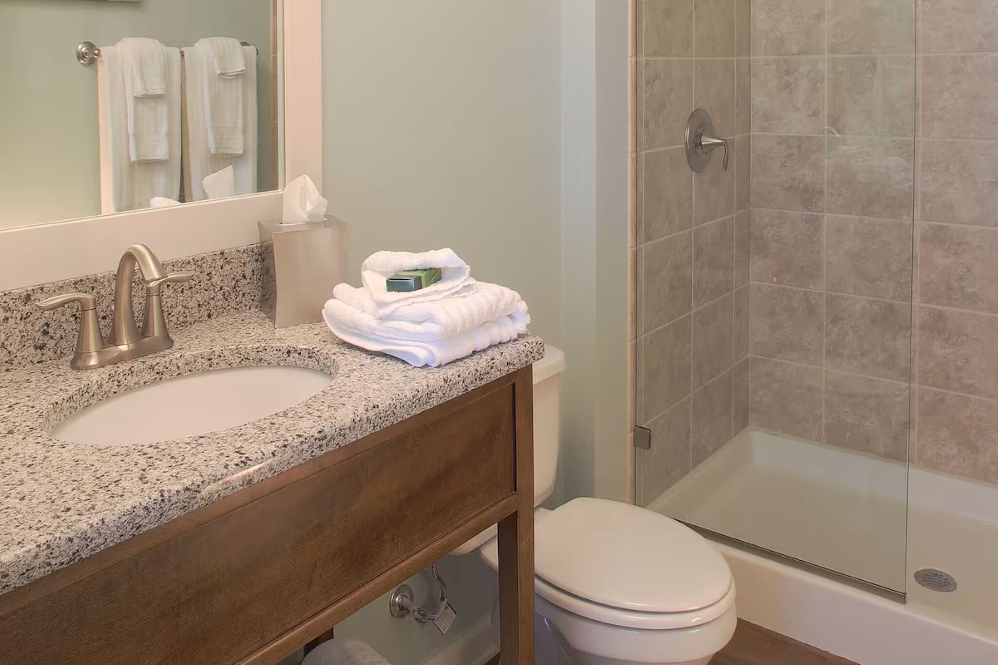 Un baño con lavamanos de encimera de granito, espejo, inodoro y ducha con mampara de vidrio.