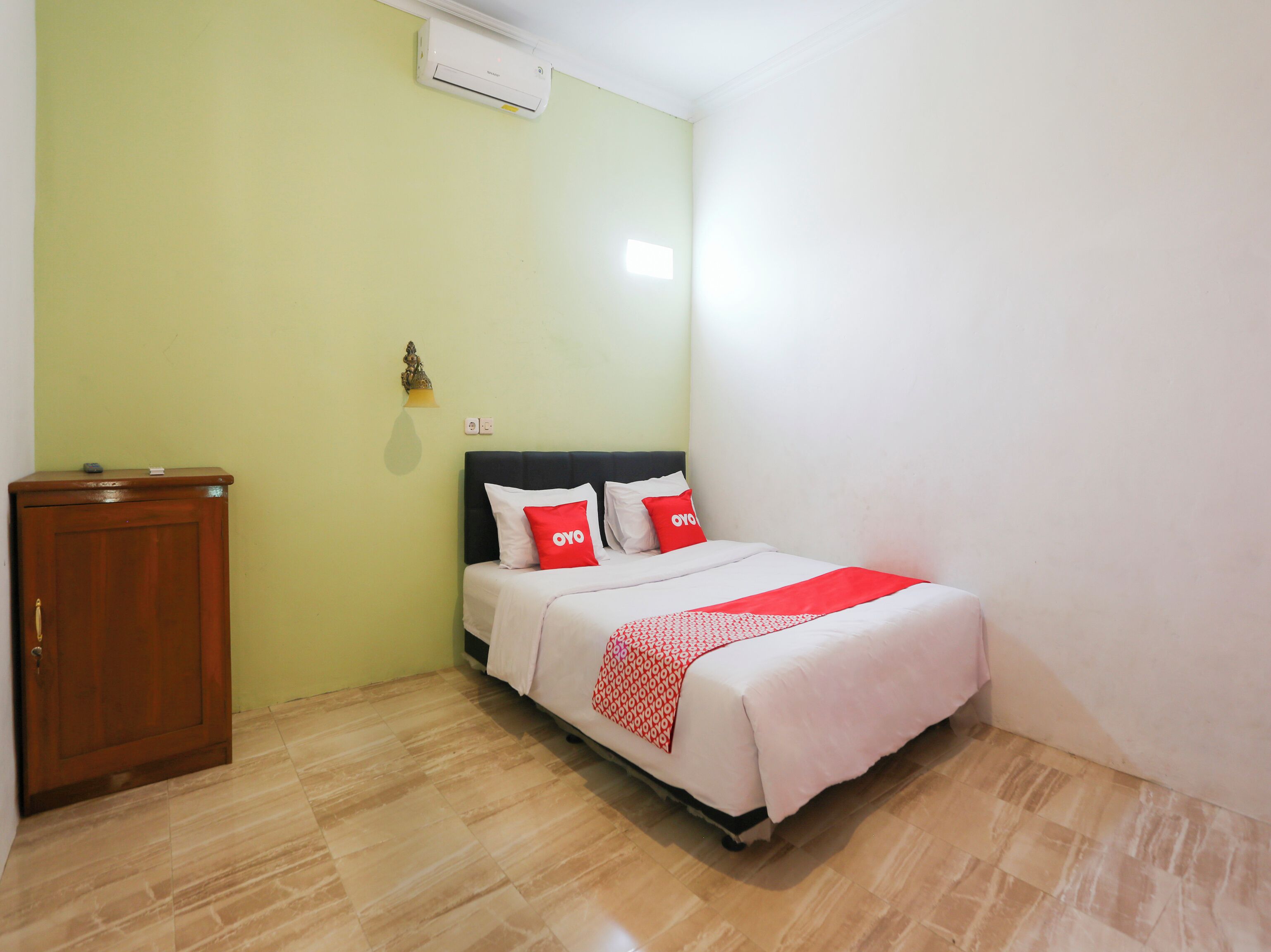 A small, neatly arranged bedroom with a bed, a wooden cabinet, and an air conditioning unit on the wall.