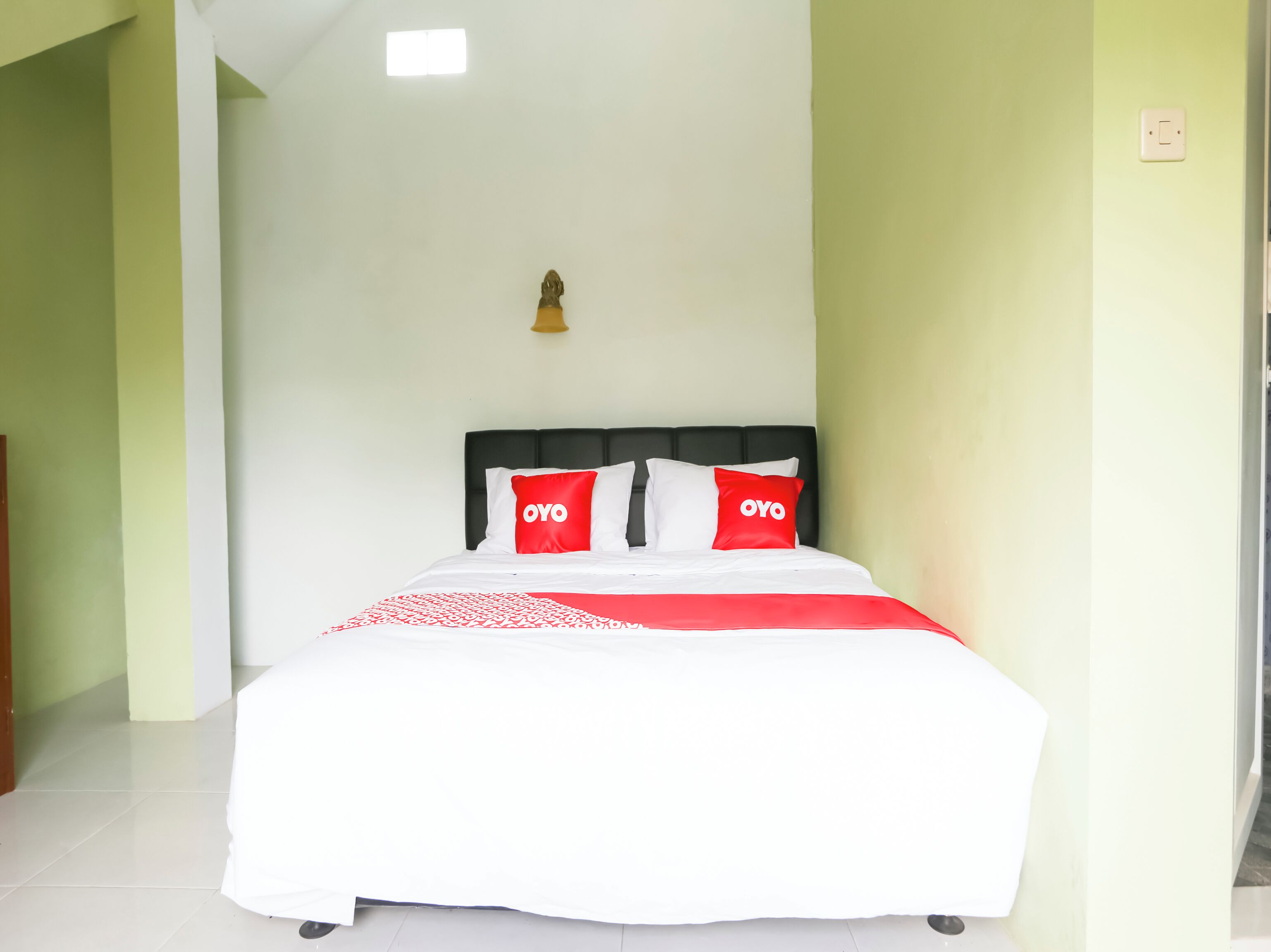 A bed with white bedding and two red pillows with "OYO" text, a black headboard, and a small wall-mounted light fixture.