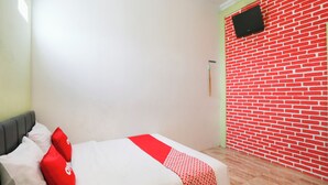 Un dormitorio con cama, almohadas, una colcha estampada en rojo y blanco, pared roja, suelo de madera y televisor montado en la pared.
