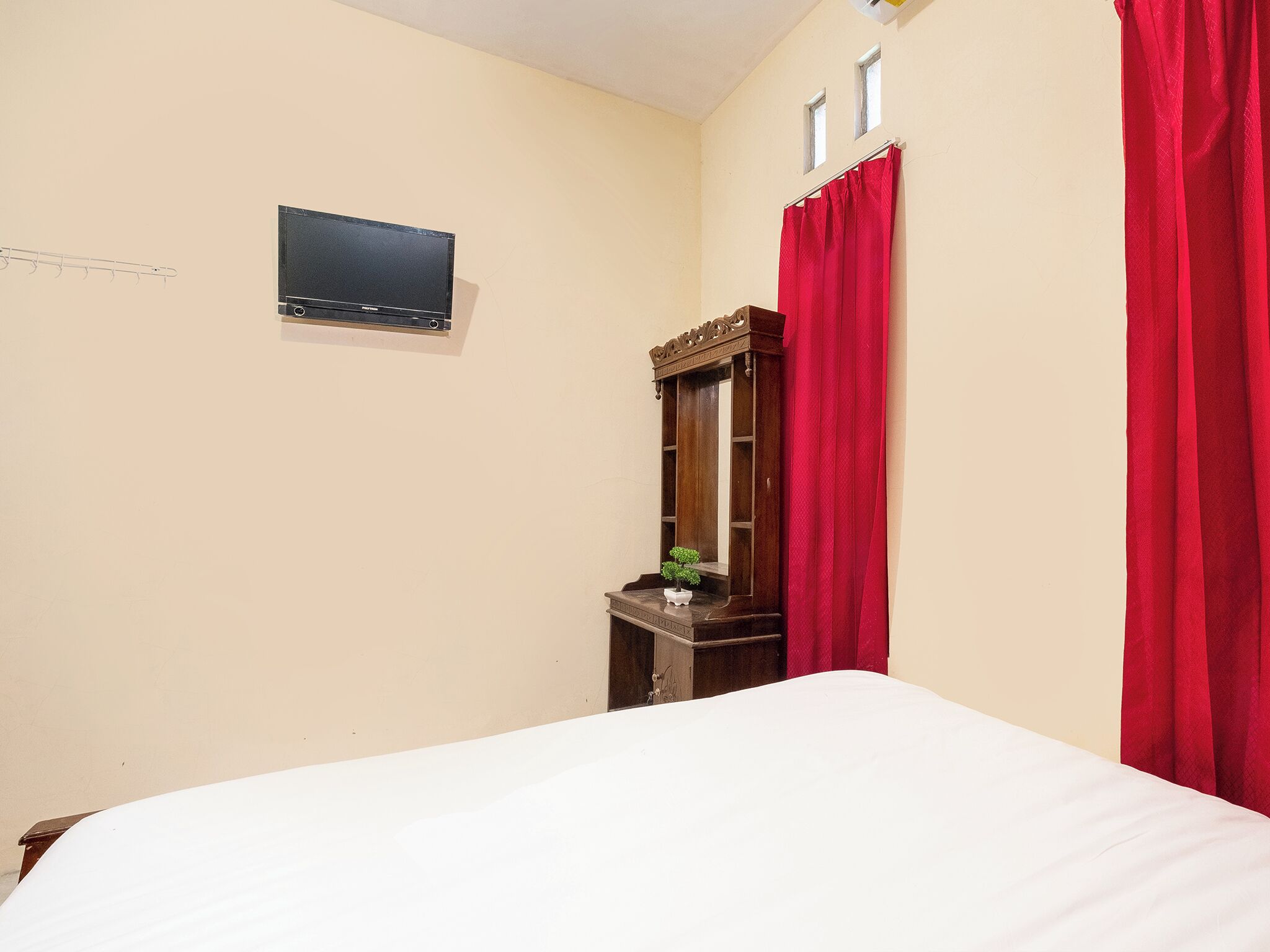 A bedroom with a bed, a TV mounted on the wall, a wooden dresser, and a red curtain.