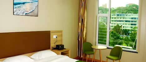 A hotel room with a bed, a desk with a lamp, two chairs, a window with a view of buildings, and a painting on the wall.