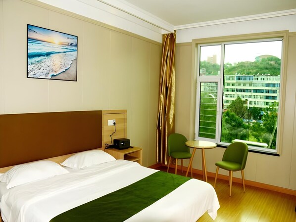 A hotel room with a bed, a desk with a lamp, two chairs, a window with a view of buildings, and a painting on the wall.