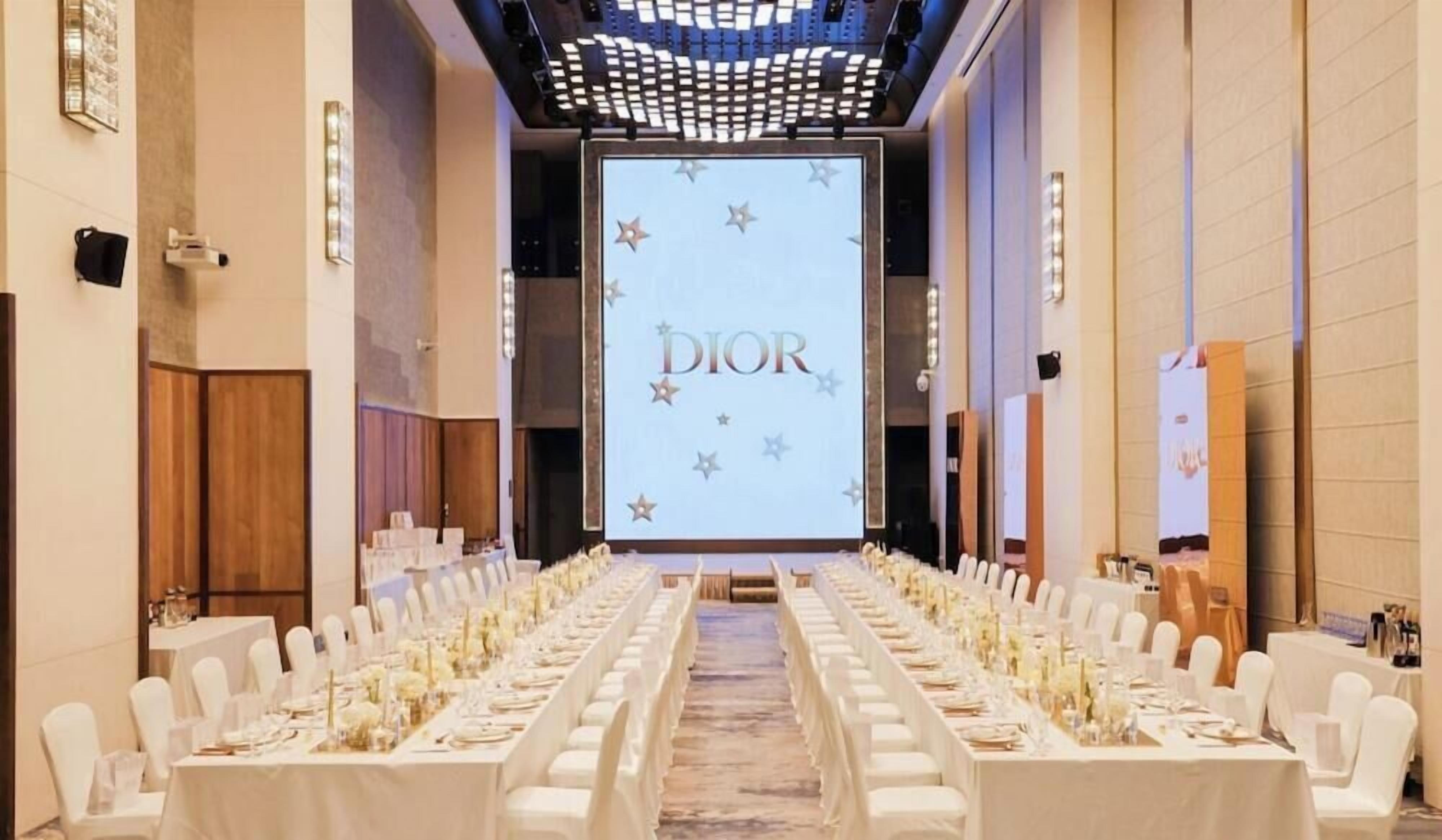 Un salón de conferencias con una larga mesa dispuesta para una reunión, una pantalla mostrando la marca 'DIOR' y un diseño de techo moderno.