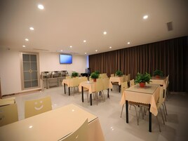 Uma zona de refeições moderna com várias mesas e cadeiras, uma TV de ecrã plano na parede e uma área de cozinha com porta de vidro.