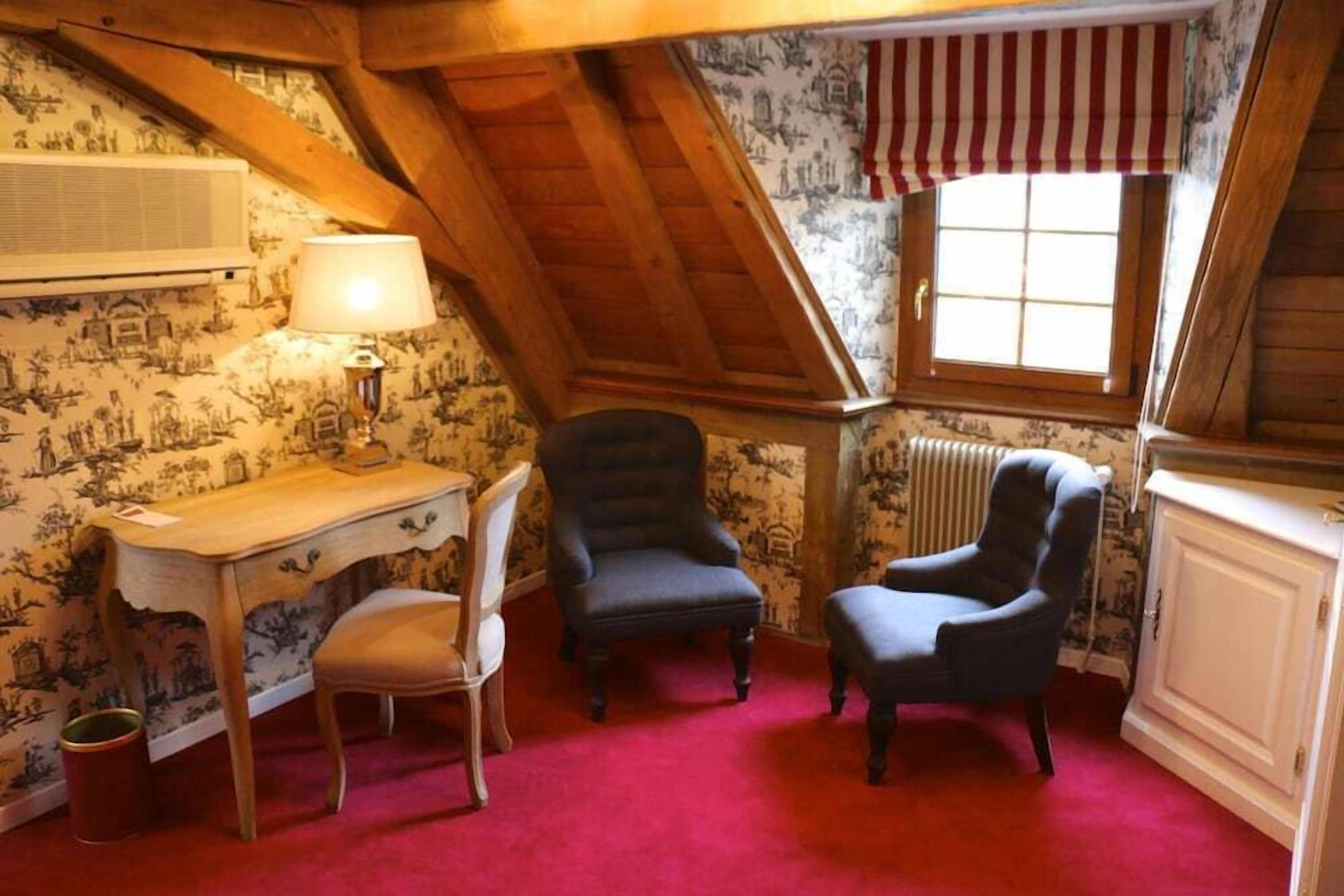 Une pièce avec un plafond en pente, un bureau avec une chaise, deux fauteuils, un radiateur et un papier peint à motifs.