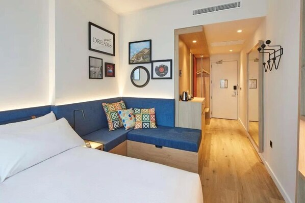 Une chambre d’hôtel moderne avec un lit, un canapé bleu et un petit coin salon. Les murs sont ornés de tableaux encadrés, le sol est en bois et un couloir mène aux autres chambres.