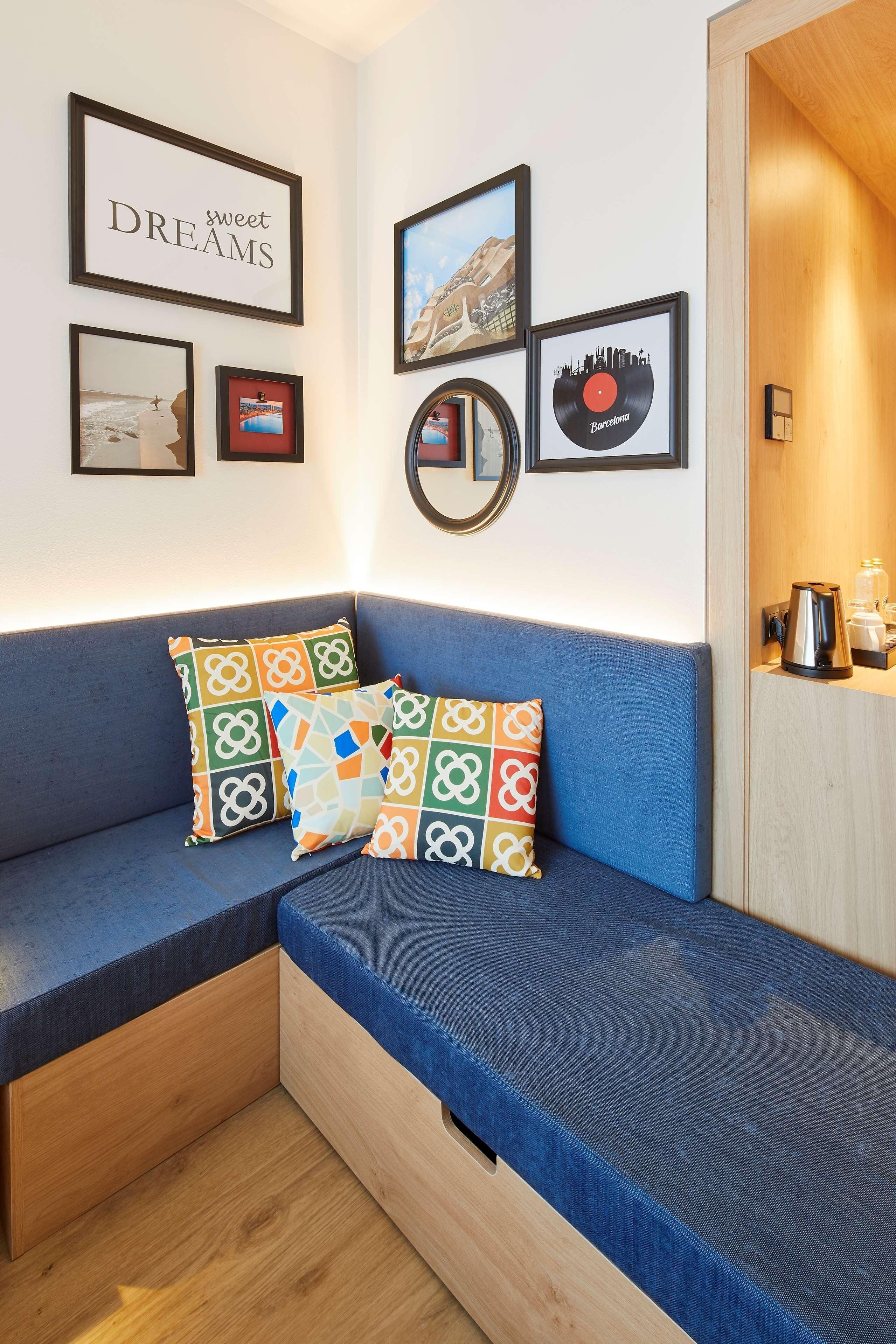 Une pièce avec un canapé bleu, des coussins colorés, un mur orné de tableaux encadrés et un meuble en bois sur lequel se trouve une bouilloire.
