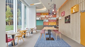 A modern dining area with round tables and chairs, a blue carpet, and a kitchen area with colorful cabinets. - Hampton By Hilton Barcelona Fira Gran Via (L'Hospitalet de Llobregat)