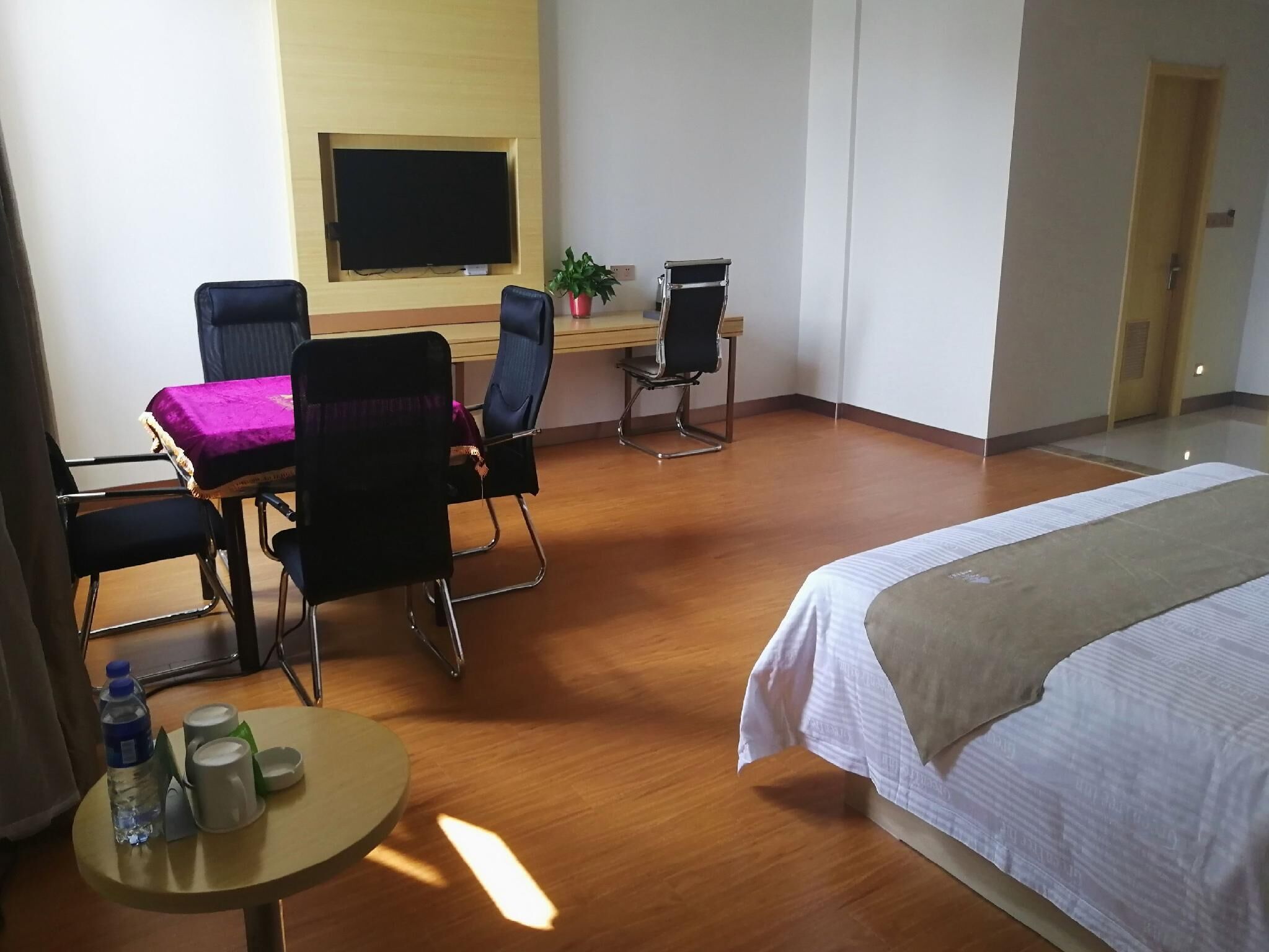 Ein Hotelzimmer mit Bett, Schreibtisch mit Stuhl, Wandfernseher sowie ein kleiner Tisch mit Wasserflasche und Bechern.
