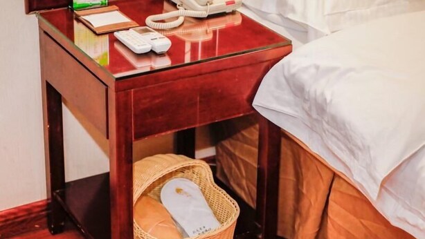 A hotel room with a wooden nightstand, a telephone, a basket with slippers, and a bed with white bedding.