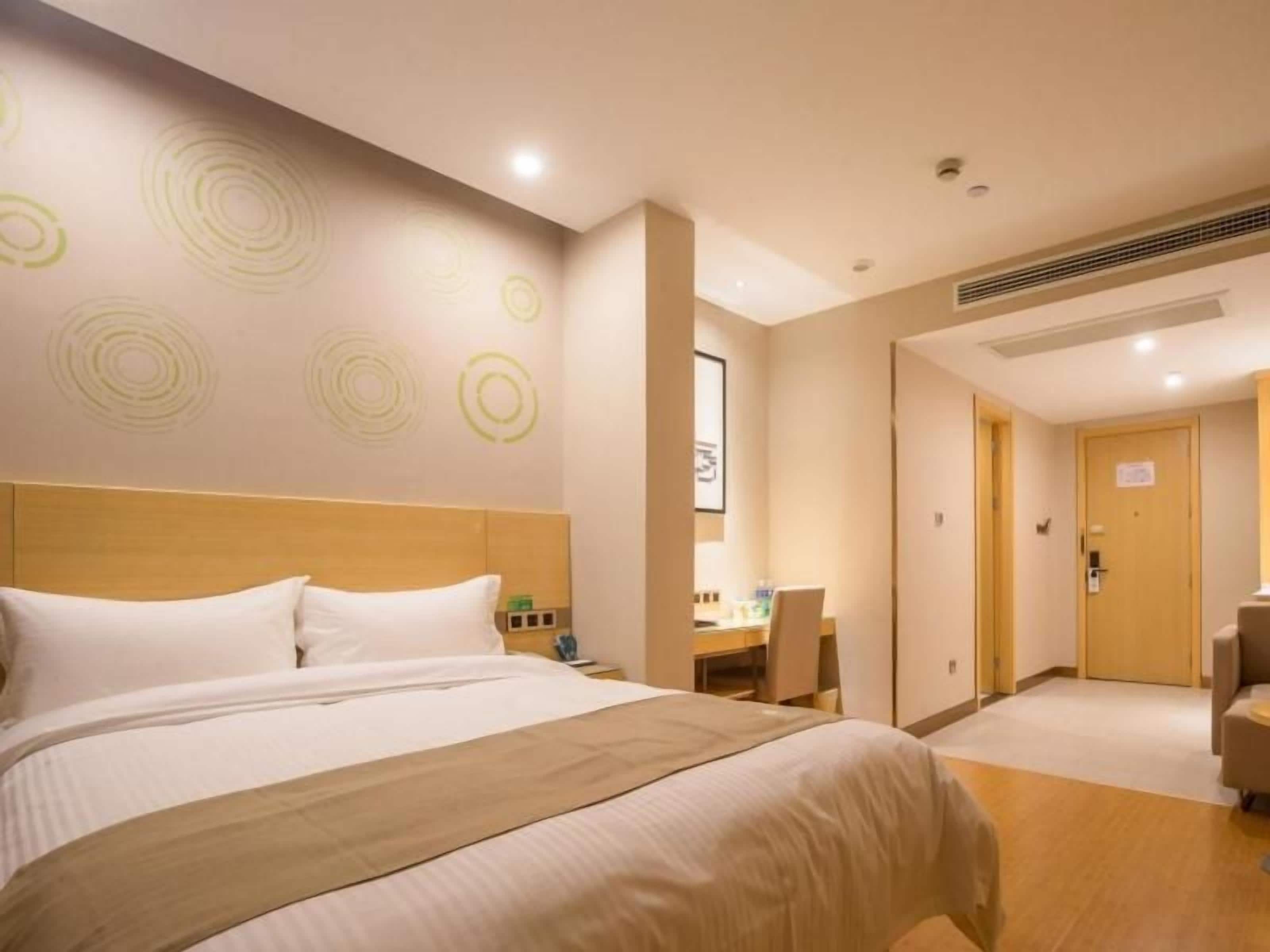 A hotel room with a large bed, a desk, and a chair. There is a door leading to another room.