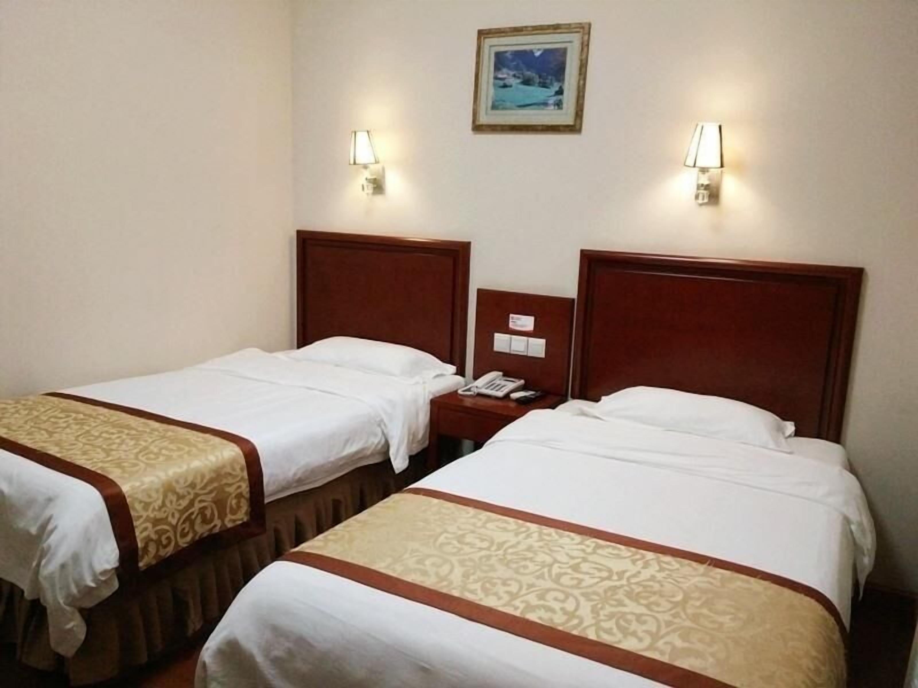 A hotel room with two single beds, each with a wooden headboard and a beige bedspread. There are two bedside tables with lamps and a small framed picture on the wall above the beds.