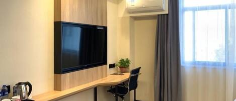 A modern hotel room with a flat-screen TV mounted on the wall, a wooden desk with a chair, a small potted plant, and a large window with curtains.