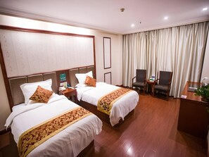 Habitación de hotel con dos camas, un escritorio, dos sillas, una planta y un ventanal con cortinas.