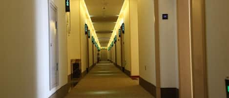 A long, narrow hotel corridor with doors on both sides, each marked with a green sign indicating room numbers.