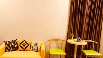 A yellow sofa with patterned cushions, two green chairs, and a small round table with a plant and a bottle.