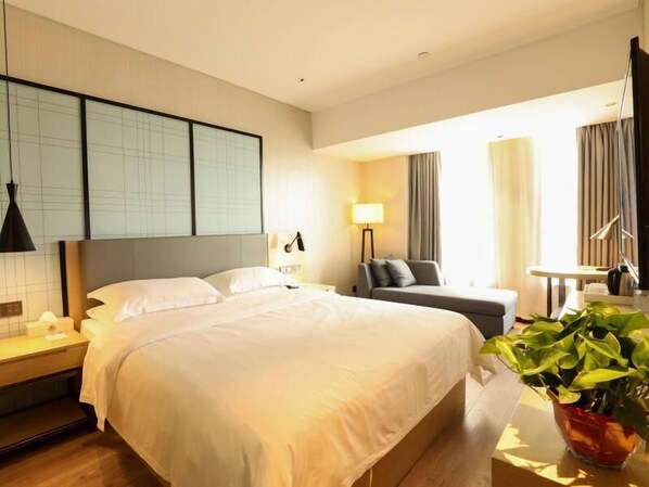 A hotel room with a large bed, a bedside table, a lamp, a sofa, a small table, and a potted plant.