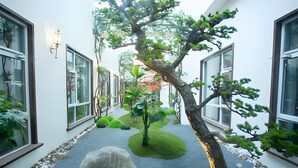 Un étroit sentier bordé de murs blancs percés de grandes fenêtres, qui mène à un petit jardin agrémenté d’un arbre et de rochers.
