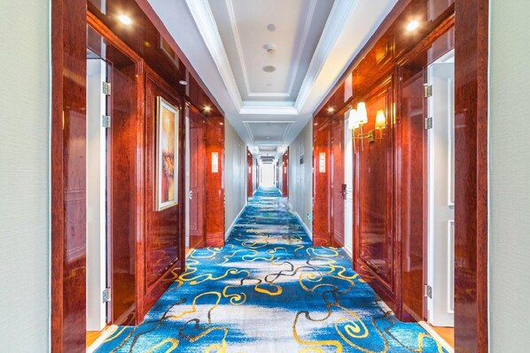 Un couloir d’hôtel avec des portes en bois, une moquette bleue ornée de motifs dorés et un éclairage encastré.