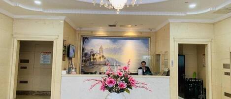 A hotel lobby with a reception desk, a chandelier, a painting on the wall, and a vase with flowers on a table.