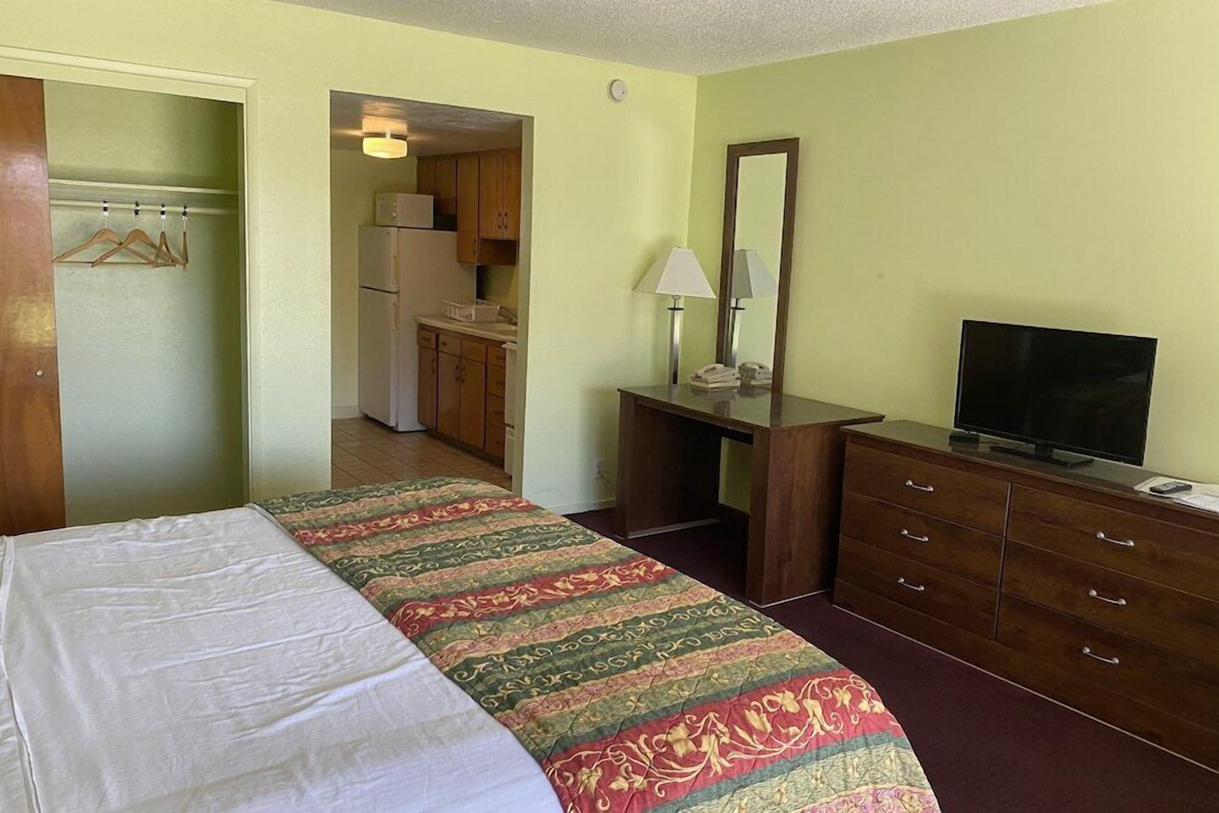 A hotel room with a bed, a desk with a lamp, a dresser, a television, and a kitchenette in the background.