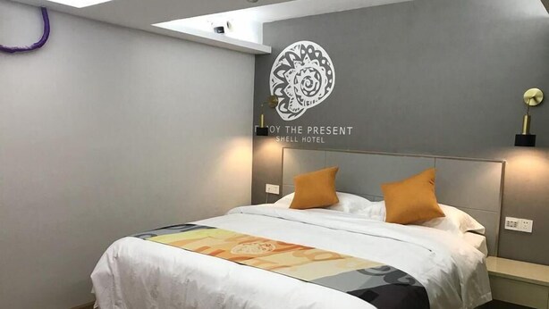 Ein Hotelzimmer mit Bett, einem an der Wand montierten Licht und einem kleinen Regal. Das Kopfteil trägt ein Logo.