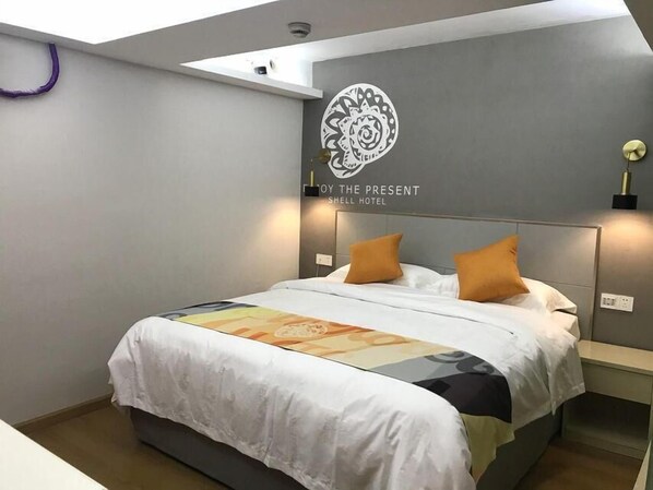 A hotel room with a bed, a headboard with a logo, a wall-mounted light, and a small shelf.