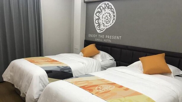 Zwei Betten mit weißen Laken und orangefarbenen Kissen, ein grauer Vorhang und eine Wand mit dem Logo und dem Schriftzug für Shell Hotel.