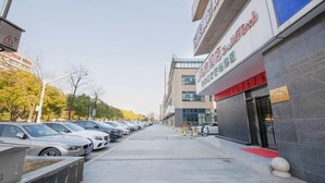 A hotel with a modern facade, a parking lot with several cars, and a clear sky. - Shell Wuxi Huishan District Wanda Plaza Yanqiao Subway Station Hotel (Wuxi)