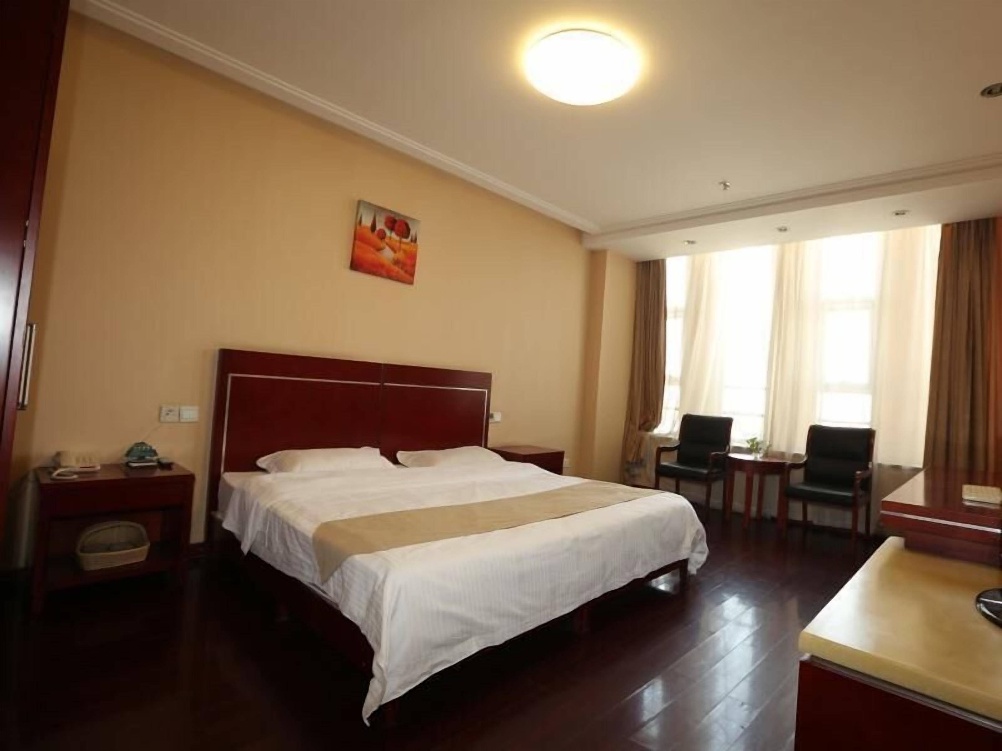 A hotel room with a large bed, a desk, two chairs, a painting on the wall, and a window with curtains.