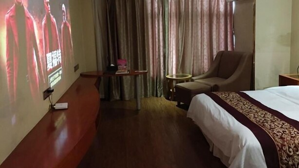 A hotel room with a bed, a seating area with a chair and a small table, a projection screen on the wall, and curtains. - Greentree Inn Wuxi Huishan High-speed Rail Qianzhou Chongwen Road Hotel (Wuxi)