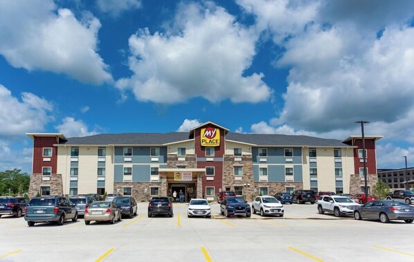 A multi-story hotel with a 'My Place' sign, a parking lot with several cars, and a clear blue sky with clouds.