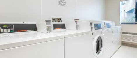 A laundry facility with multiple washing machines and dryers, a control panel, and a window with blinds.