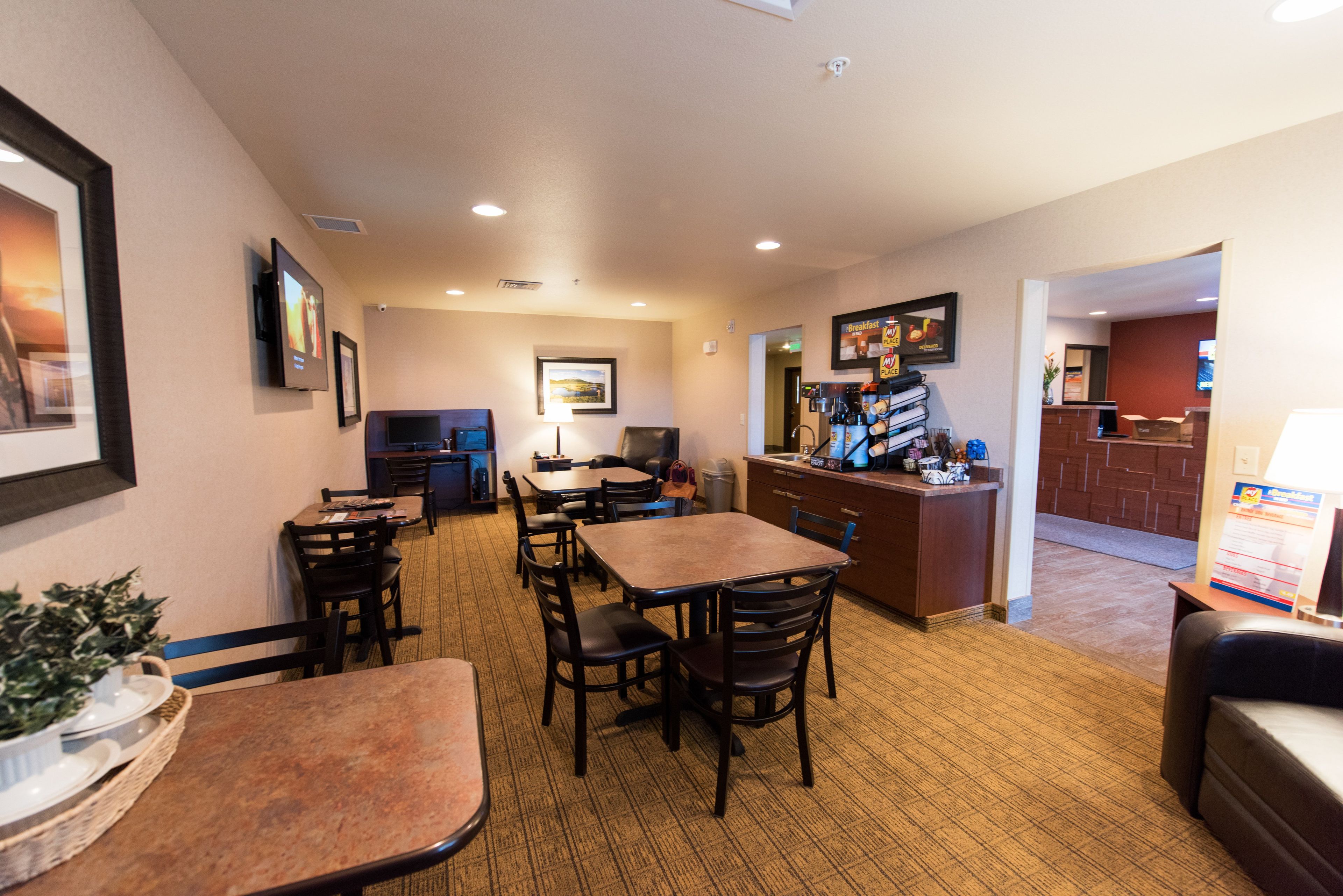 A hotel dining area with tables and chairs, a coffee station, and a television mounted on the wall.