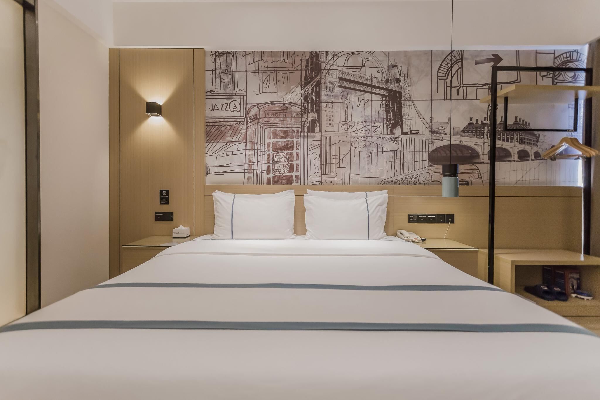 Hotellihuone, jossa on suuri sänky, seinämaalaus, yöpöytä lampun kanssa ja hylly, jossa on kenkiä.