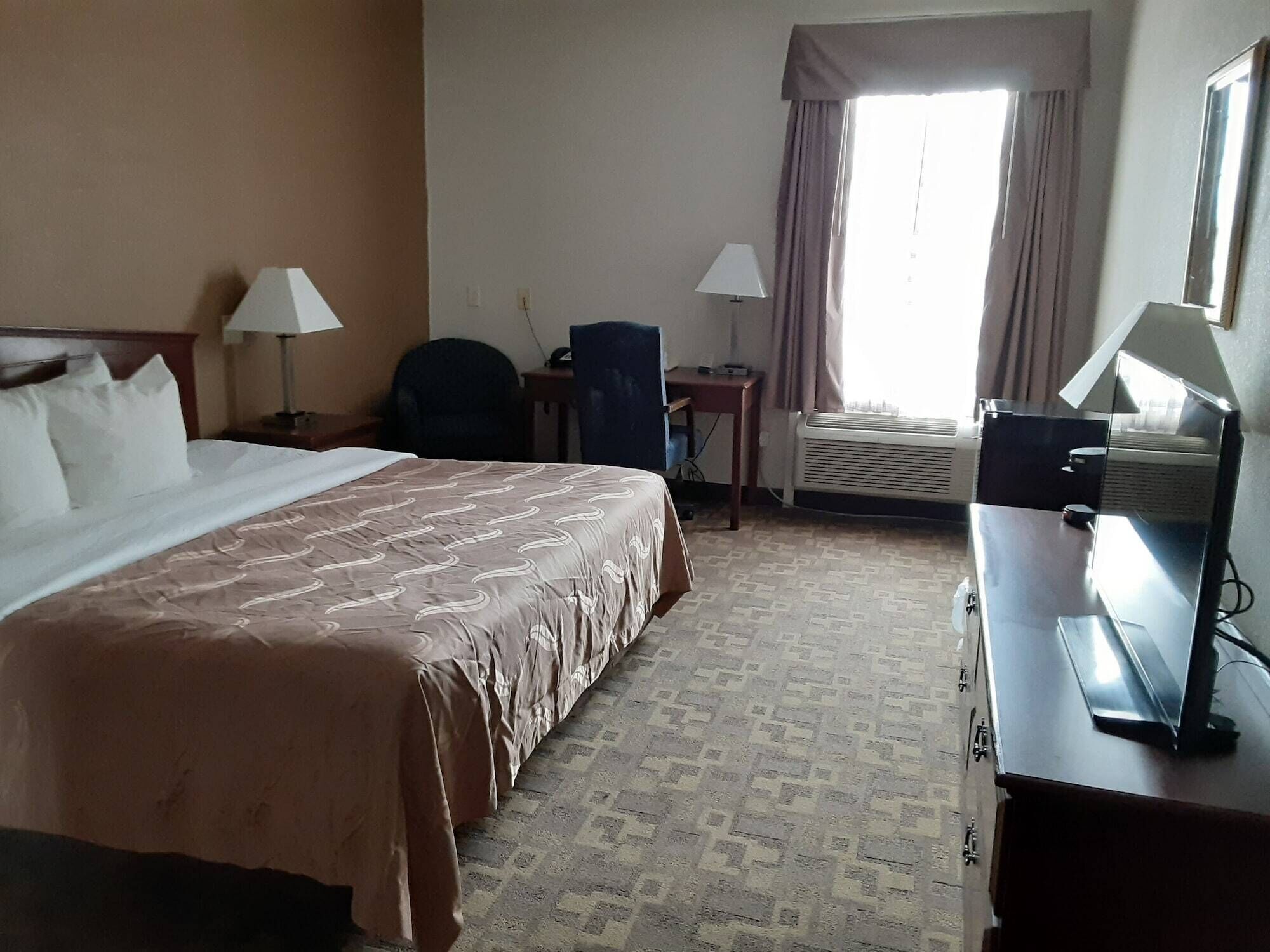 A hotel room with a bed, two bedside lamps, a desk with a chair, an air conditioning unit, and a television.