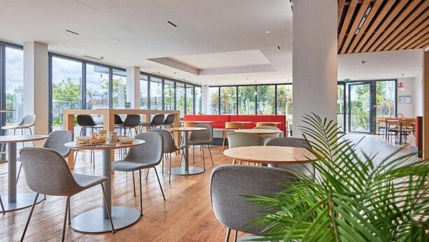 A modern indoor space with round tables and chairs, a red seating area, and large windows offering a view of greenery outside. - B&B Hotel Deauville-touques (Touques)