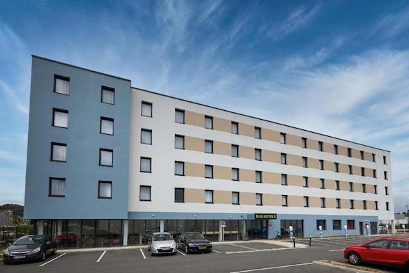 A modern hotel building with a flat facade, multiple windows, and a parking lot with several cars. - B&B Hotel Deauville-touques (Touques)