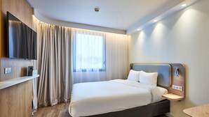 A modern hotel room with a large bed, a flat-screen TV mounted on the wall, a bedside table, and a window with curtains. - B&B Hotel Deauville-touques (Touques)