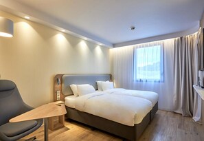 Ett hotellrum med en säng, en stol, ett skrivbord och ett fönster med gardiner.