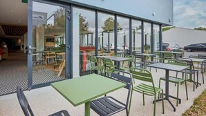 An outdoor seating area with tables and chairs, a glass door entrance, and a view of parked cars and trees. - B&B Hotel Deauville-touques (Touques)