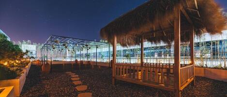 A night-time outdoor seating area with a thatched-roof structure, a glass-enclosed garden, and a pathway of stepping stones.