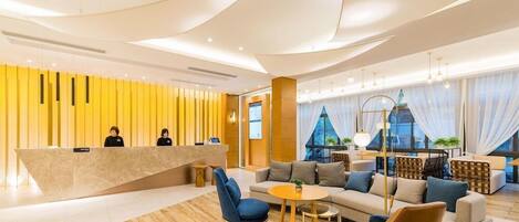 A modern hotel lobby with a reception desk, seating area, and large windows.
